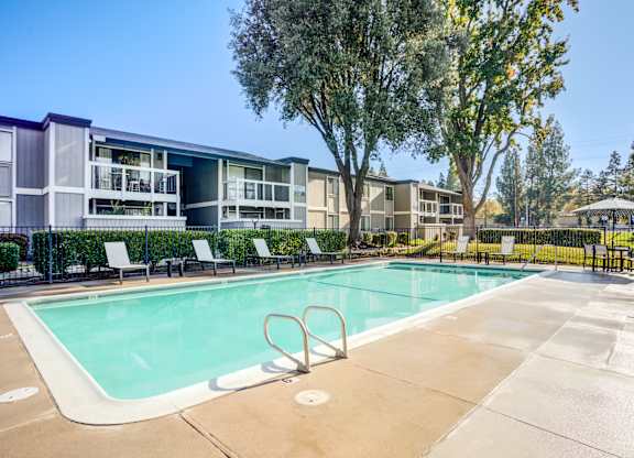 A swimming pool surrounded by trees and chairs in front of apartment buildings.