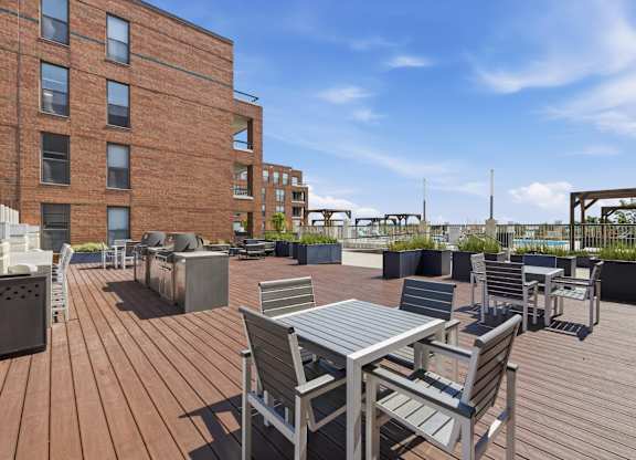 A rooftop wooden deck with metal tables and chairs in front of a grilling statioin.