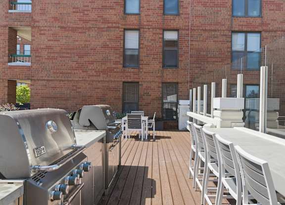 A rooftop patio with a grill and chairs is in front of a brick building.