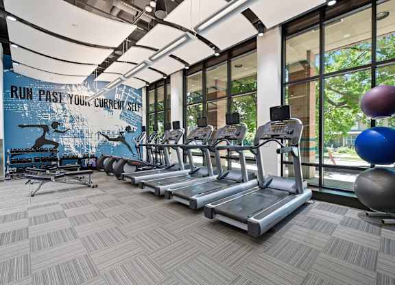 A gym with treadmills and a motivational poster on the wall in front of floor to ceiling windows.