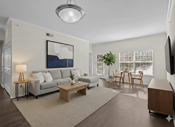 A living room with a grey couch, a coffee table, a lamp, a painting, a dining table with chairs, and a flat-screen TV mounted on the wall.