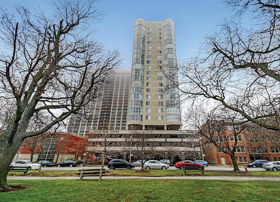 A tall building with many windows is surrounded by trees and cars.