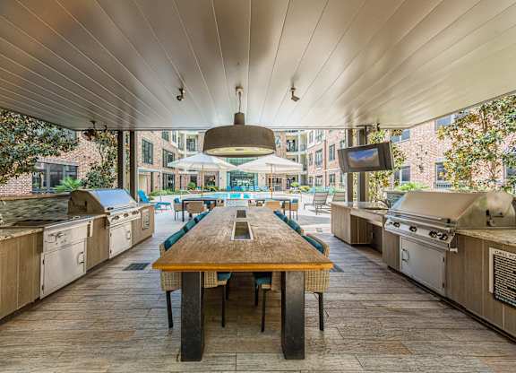 A long wooden table is in the middle of a patio with barbecue grills on either side.