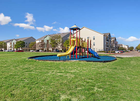 A playground with a slide and a blue mat in the middle of a grassy area.