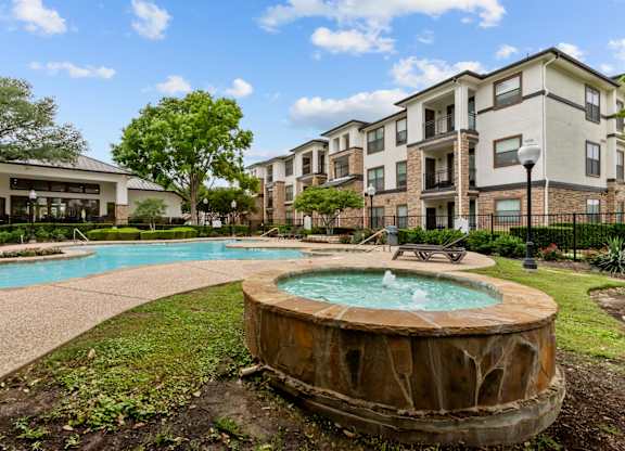 A large outdoor swimming pool with a hot tub in the middle of a grassy area in front of apartment buildings.