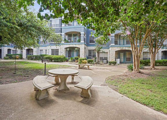 A concrete table and chairs are in the foreground of a courtyard with a building in the background.