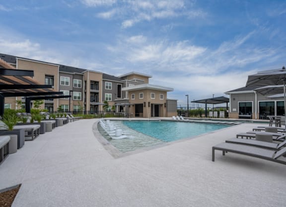 A swimming pool surrounded by sun loungers and umbrellas in front of apartment buildings.