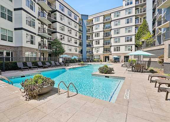 A large swimming pool surrounded by lounge chairs and umbrellas in a courtyard with apartment buildings in the background.