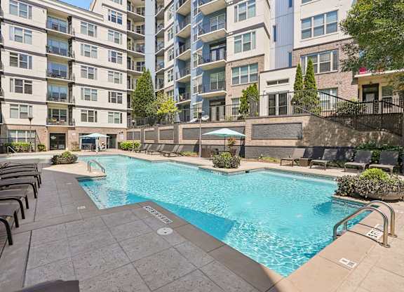A swimming pool surrounded by lounge chairs and umbrellas in front of apartment buildings.