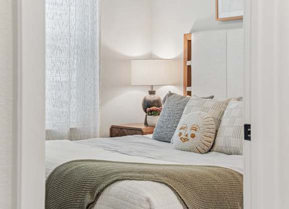 A bedroom with a bed, a nightstand with a lamp, and a window with curtains.