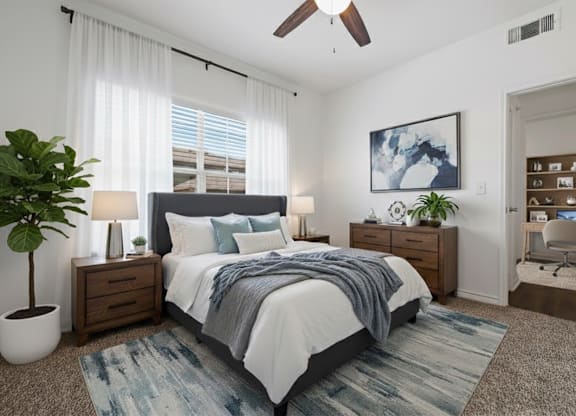 A bedroom with a bed, a plant, a ceiling fan, and a painting on the wall.