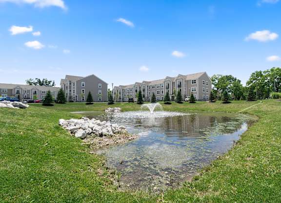 A pond with a fountain in the middle of a grassy area in front of apartment buildings.