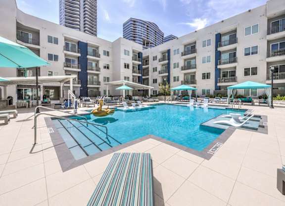 A large swimming pool with a diving board and sun loungers in front of apartment buildings.