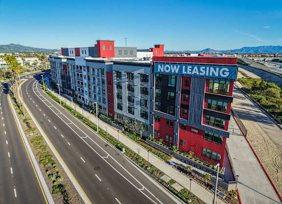 A multi-story building with a red and white exterior and a sign that reads "NOW LEASING" is surrounded by a highway.
