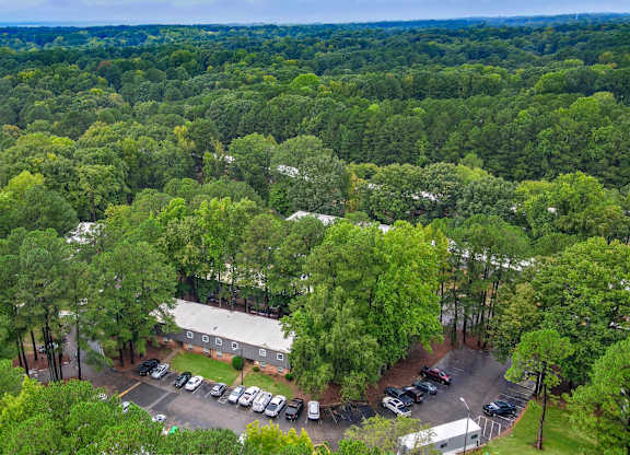 A parking lot with cars and a building surrounded by trees. at Arbors at Cary, Cary, 27511