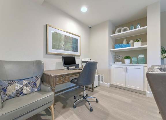 A room with a grey couch, a wooden desk with a computer, a grey chair, and a cabinet with shelves containing various items.