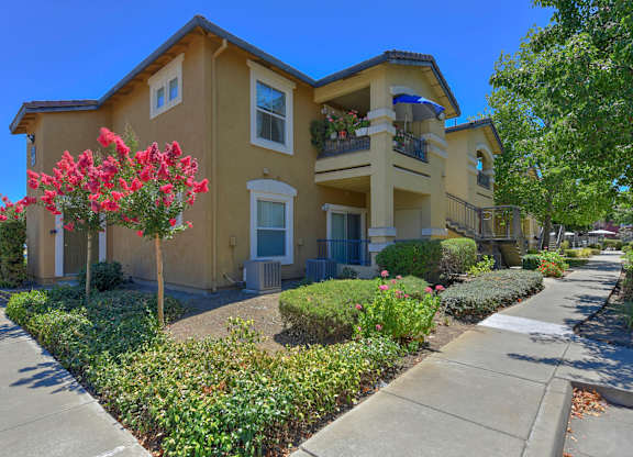 A yellow two-story house with a balcony and a flower bed in front at Hurley Creek Apartment Homes, Sacramento