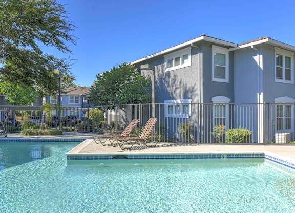 A swimming pool in front of a grey house with a fence around it.