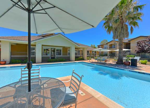 A pool with a table and chairs and an umbrella in the foreground with apartment buildings in the background at Hurley Creek Apartment Homes, Sacramento, California