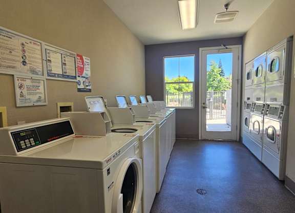 A laundromat with washers and dryers and a view of the outside through the window.
