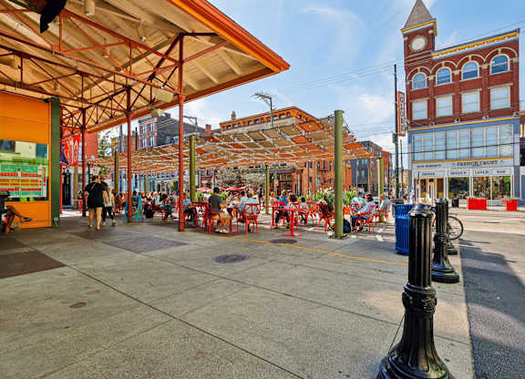 People enjoying outdoor dining at Findlay Market in Over-the-Rhine Cincinnati with red patio seating and lively atmosphere.