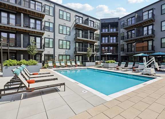 A swimming pool surrounded by lounge chairs and umbrellas in front of apartment buildings.