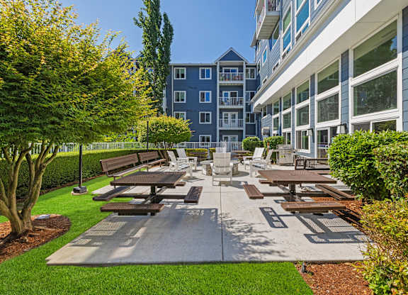 A large outdoor picnic area with benches and tables is surrounded by greenery and a building.