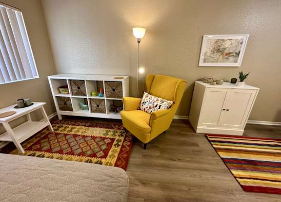 A yellow chair with a white cushion is in the corner of a room with a white shelf and a lamp.