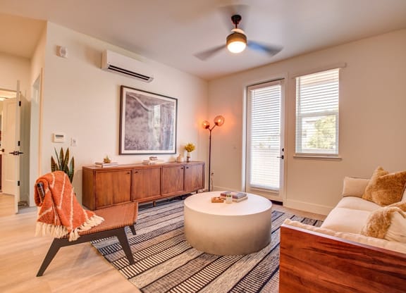 A Large Living Room With Wood-Style Plank Flooring, Area Rug, A Couch, A Chair, A Round Coffeetable, A Ceiling Fan, AC Unit, A Window With Blinds, And A Door Leading Into Patio Or Balcony Area at 38° 