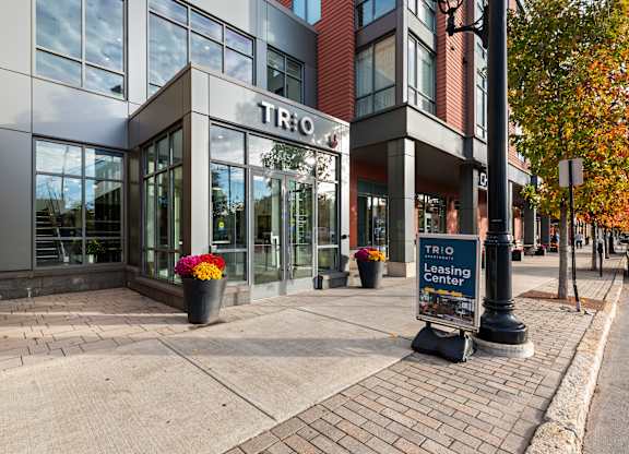 A brick building with a sign that says TRIO Leasing Center at Trio Newton Apartments, Newton, 02460