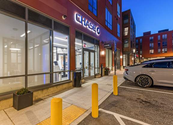A Chase bank is lit up at night with a car parked in front at Trio Newton Apartments, Massachusetts, 02460