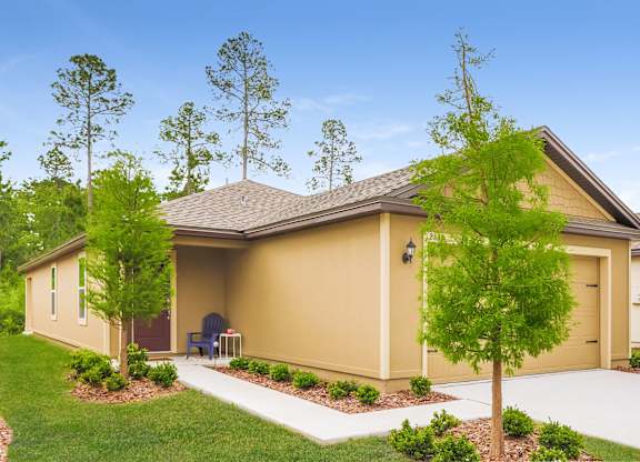 Exterior view of a single-story beige house with a brown roof, garage, manicured lawn, and blue Adirondack chair on the porch.