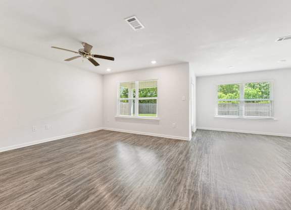 Empty room with gray wood-look flooring, white walls, ceiling fan, and two windows with blinds.