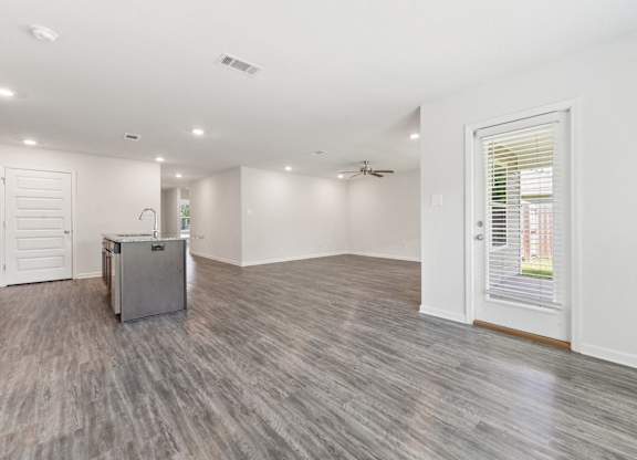 Bright, open concept living space with gray wood-look flooring, kitchen island, and backyard view through a door with blinds.