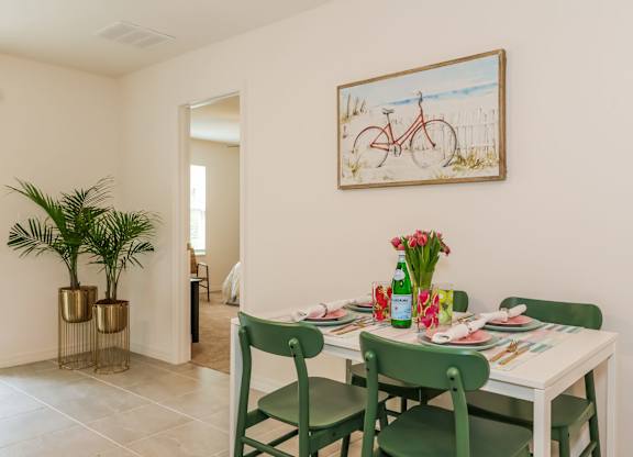 Dining room with a white table set for four, green chairs, tropical plants, and beach-themed artwork.