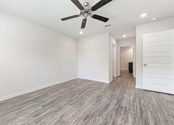Empty bedroom with gray wood-look flooring, white walls, ceiling fan, and bathroom doorway.