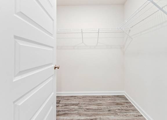 Empty walk-in closet with white walls, wood-look flooring, and wire shelving, viewed through an open door.