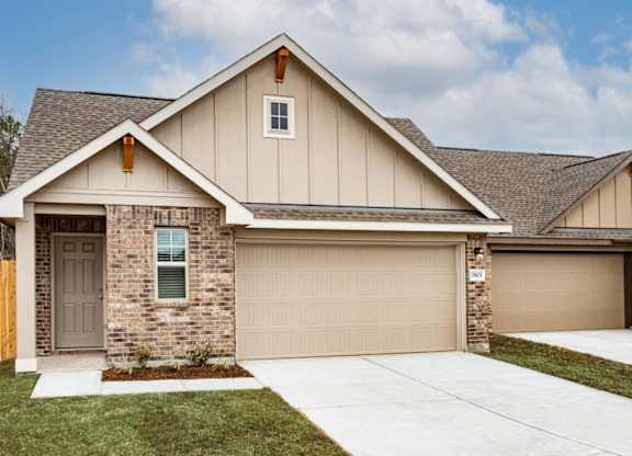 Pet-Friendly Apartments in Humble, TX - Edgewater at the Lagoon - Property Exterior with Brown and Tan Walls, White Trim, Stone Details, Parking Garage, Beige Garage Door, Plant Bed, Grass Lawns, and Concrete Driveway.
