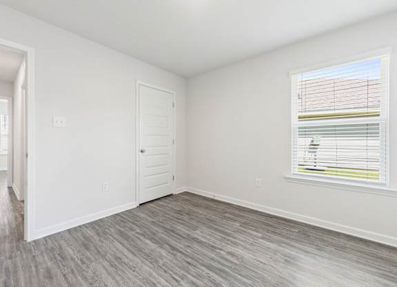 Empty bedroom with gray wood-look flooring, white walls, a window with blinds, and a white door.