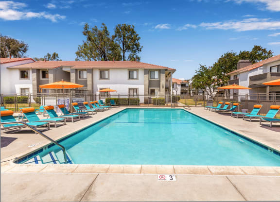 Pet-Friendly Apartments in Moreno Valley, CA - Sorelle Apartments - Classic Rectangular Swimming Pool Flanked by Rows of Blue Lounge Chairs and Orange Umbrellas with Views of the Apartment Homes