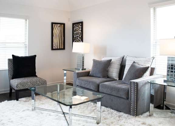 Two-Bedroom Apartments in Humble, TX - Edgewater at the Lagoon - Living Room with Vinyl Plank Flooring, Fluffy White Area Rug, Glass Cofee Table, End Tables with Lamps, Grey Couch, Graphic Cushioned Chair, Two Windows, and Framed Wall Art.