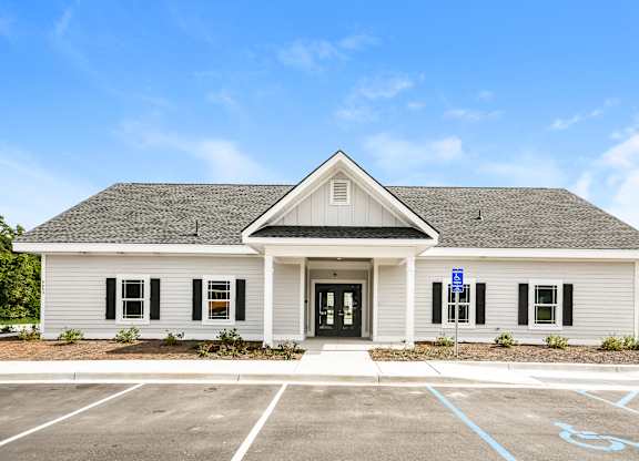 A small white building with a black door and windows, a sign on the door, and a parking lot in front.