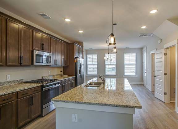 Houses for Rent in North Charleston, SC - Sumner Village - Kitchen with Wood-Style Flooring, Granite Countertops, Stainless Steel Appliances Including a Refrigerator, Oven, and Built-In Microwave, Brown Cabinetry, a Large Kitchen Island, Overhead Hanging Lights, and a View of the Connected Living Area.