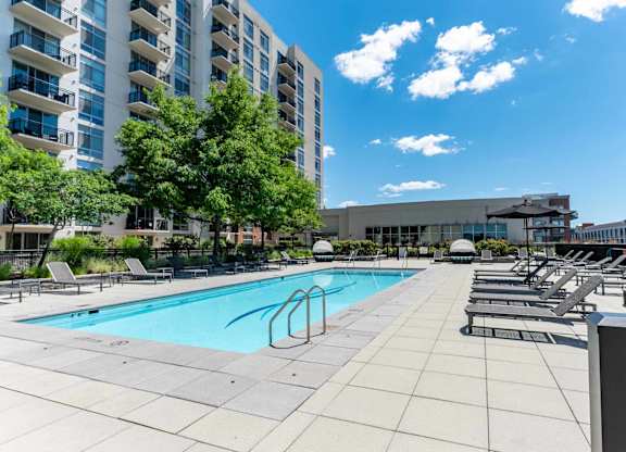 A swimming pool surrounded by lounge chairs and trees in front of a building.