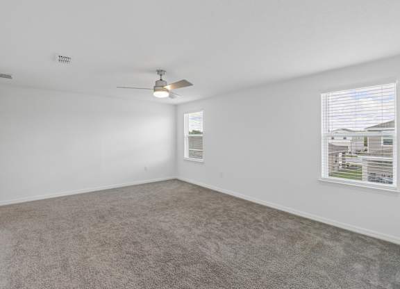 A room with a carpeted floor, a ceiling fan, and a window showing a view of buildings outside.