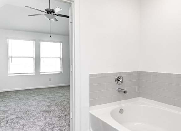 Humble, TX, Apartments for Rent - Edgewater at the Lagoon - Attached Bathroom with Bath Tub, Grey Tile Backsplash, and View of Bedroom with Carpeted Flooring and Ceiling Fan.