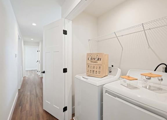 Laundry area with washer, dryer, shelving, and wood-style flooring in a bright, modern hallway.