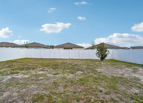 A tree stands alone in a grassy field with a white fence and houses in the background.