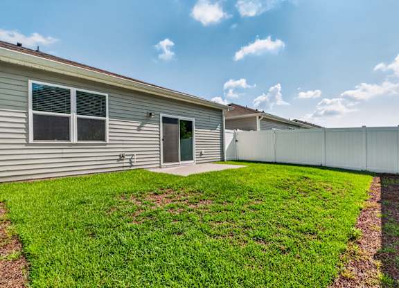 A house with a grey siding and a green door is surrounded by a white fence and has a green lawn.