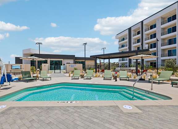A large outdoor swimming pool with lounge chairs and umbrellas in front of a modern building.